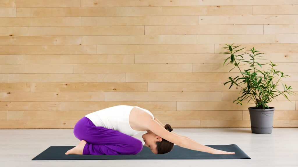 woman doing the Child’s Pose on a black yoga mat indoors during her morning stretching